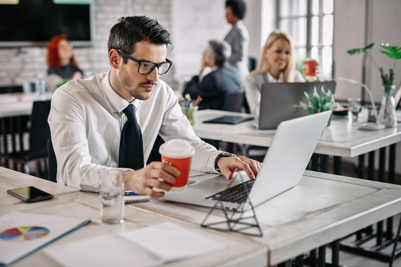 young-businessman-sitting-office-working-laptop-while-drinking-coffee-there-are-people-background